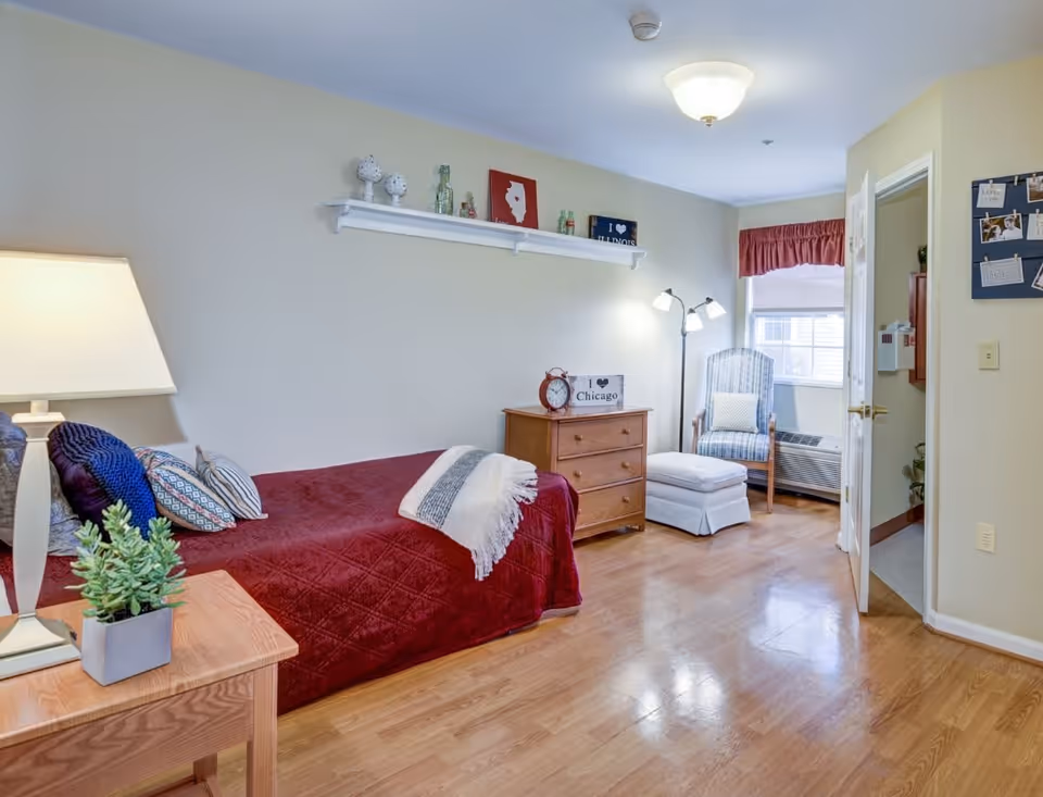 A cozy bedroom with a single bed covered in a red quilt and multiple pillows. A wooden nightstand with a lamp and a small plant is beside the bed. Across from the bed is a wooden dresser with a clock and a sign that says 'I love Chicago'. A comfortable armchair with an ottoman is placed near a window with a red valance. The room has light-colored walls, wooden flooring, and a ceiling light fixture.