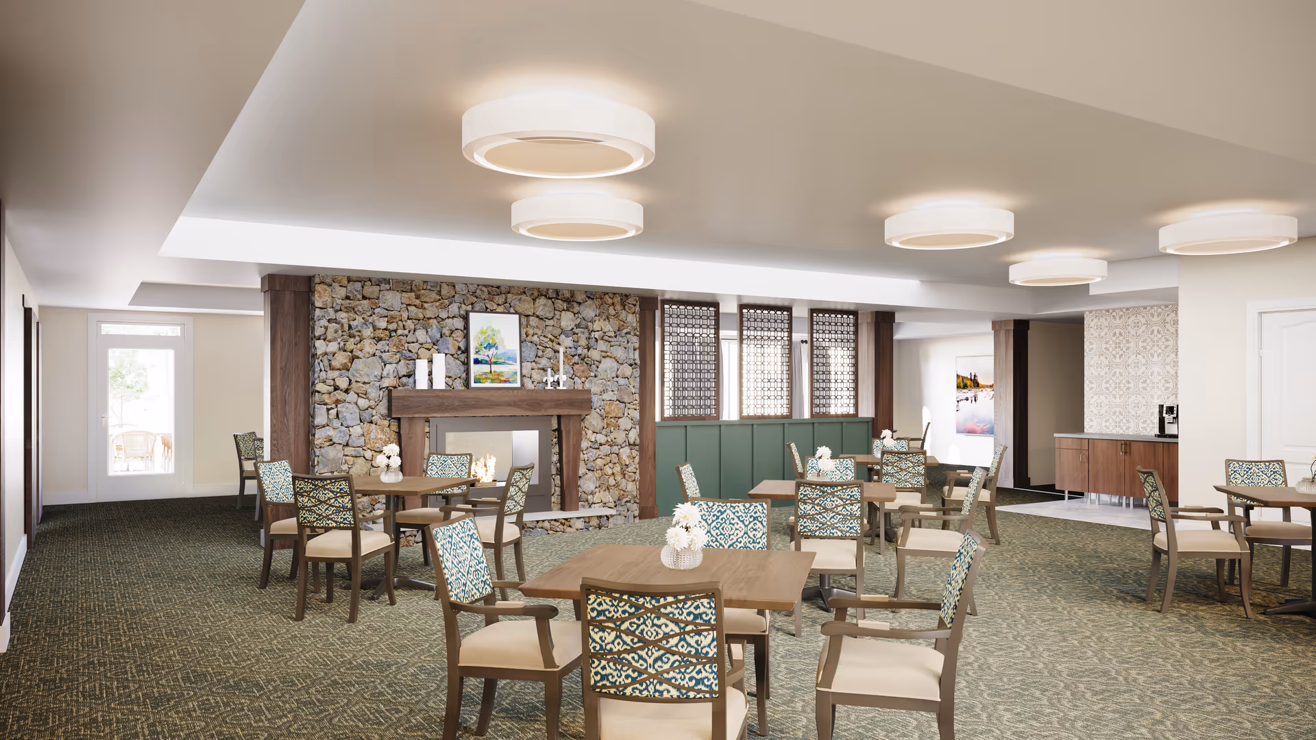 A spacious dining room with multiple wooden tables and cushioned chairs featuring patterned upholstery. The room has a stone fireplace with decorative items and a framed picture above it. The ceiling has modern circular light fixtures, and there is a sideboard with a coffee machine in the background. The room is carpeted with a green patterned carpet and has large windows with decorative wooden panels.