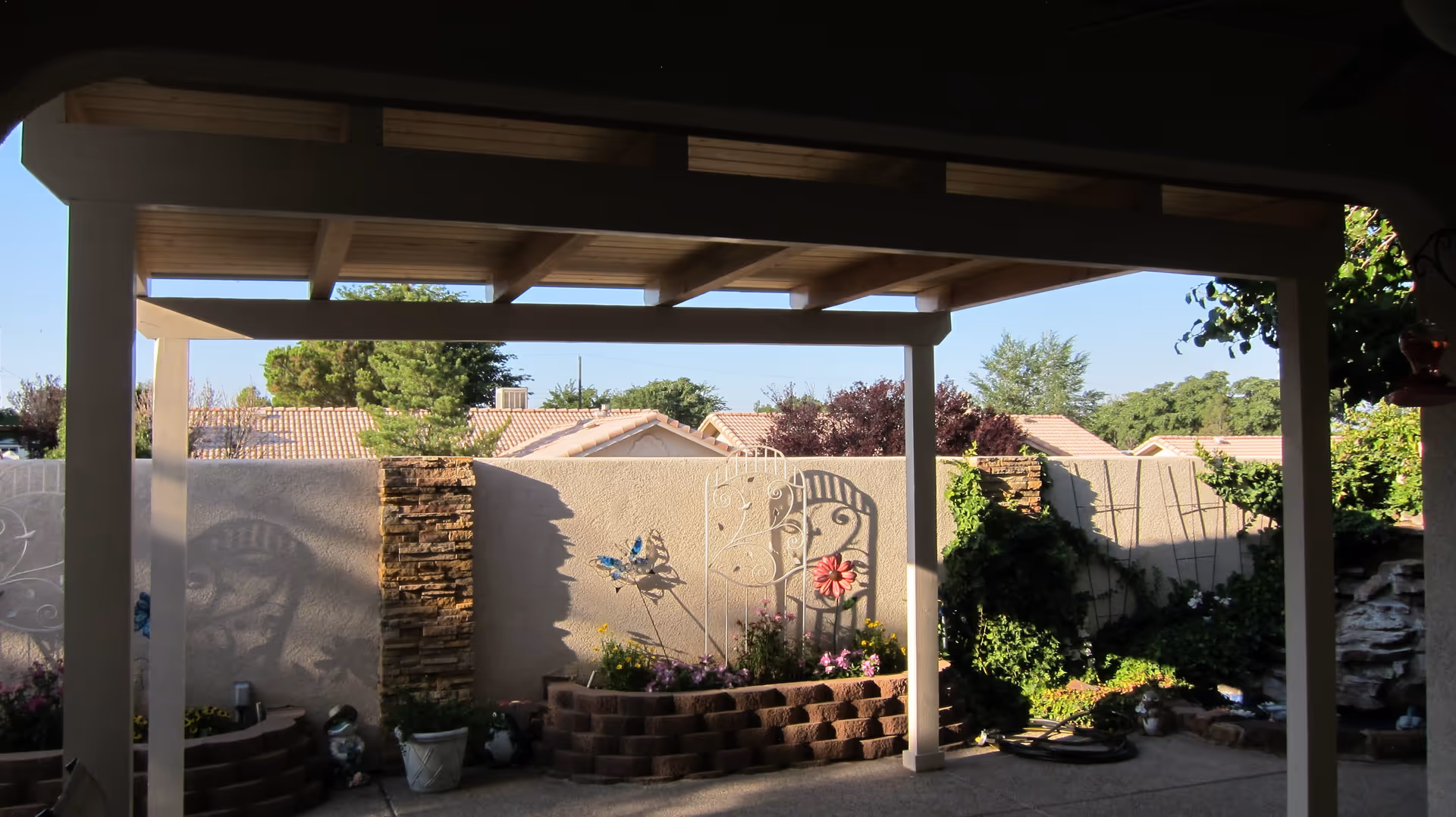 View of a covered patio area with wooden beams overhead, overlooking a garden with flower beds, decorative metal trellises, and a stone water feature. There is a beige stucco wall with stone accents and greenery climbing on trellises, with rooftops and trees visible beyond the wall under a clear blue sky.