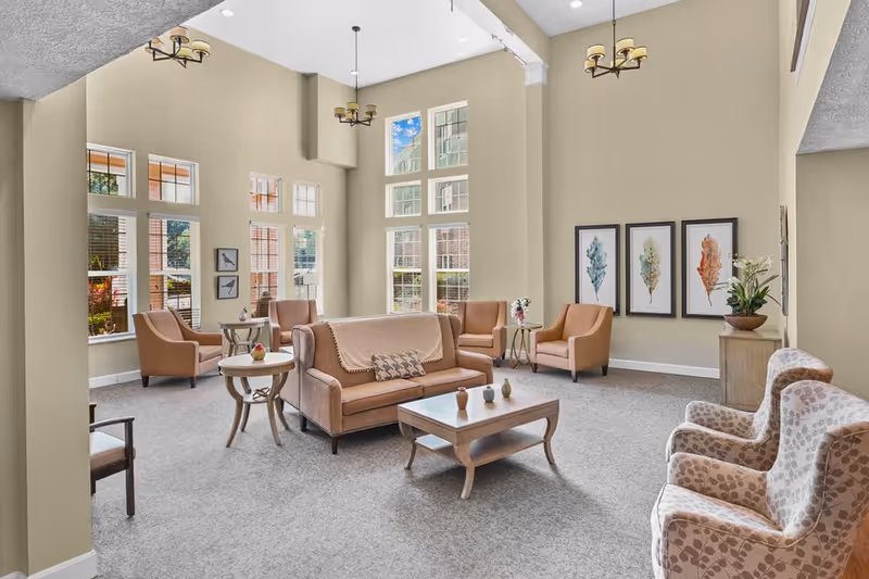A bright and spacious living room area with high ceilings and large windows letting in natural light. The room features beige walls and carpeted floors, with several comfortable armchairs and a sofa arranged around a wooden coffee table. Decorative framed artwork and plants add a cozy touch to the space.