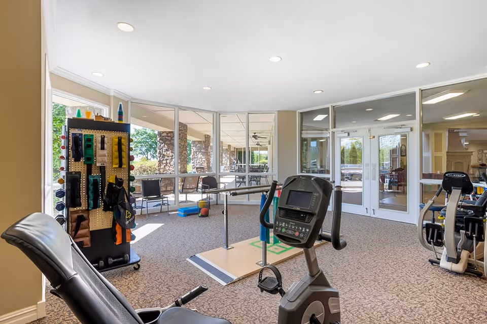 A bright exercise room with large windows showing an outdoor patio area. The room contains various fitness equipment including a stationary bike, a recumbent bike, and a rack with resistance bands and other exercise accessories. The room has carpeted flooring and glass doors leading to another interior space.
