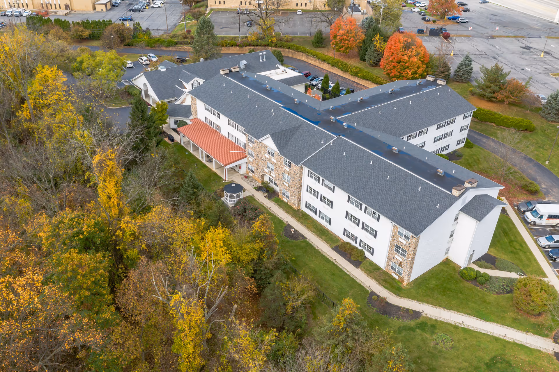 Aerial view of a senior living facility building surrounded by trees with autumn foliage and adjacent parking areas. The building has a gray roof, white walls, and a stone facade section with a covered entrance and a small gazebo nearby.