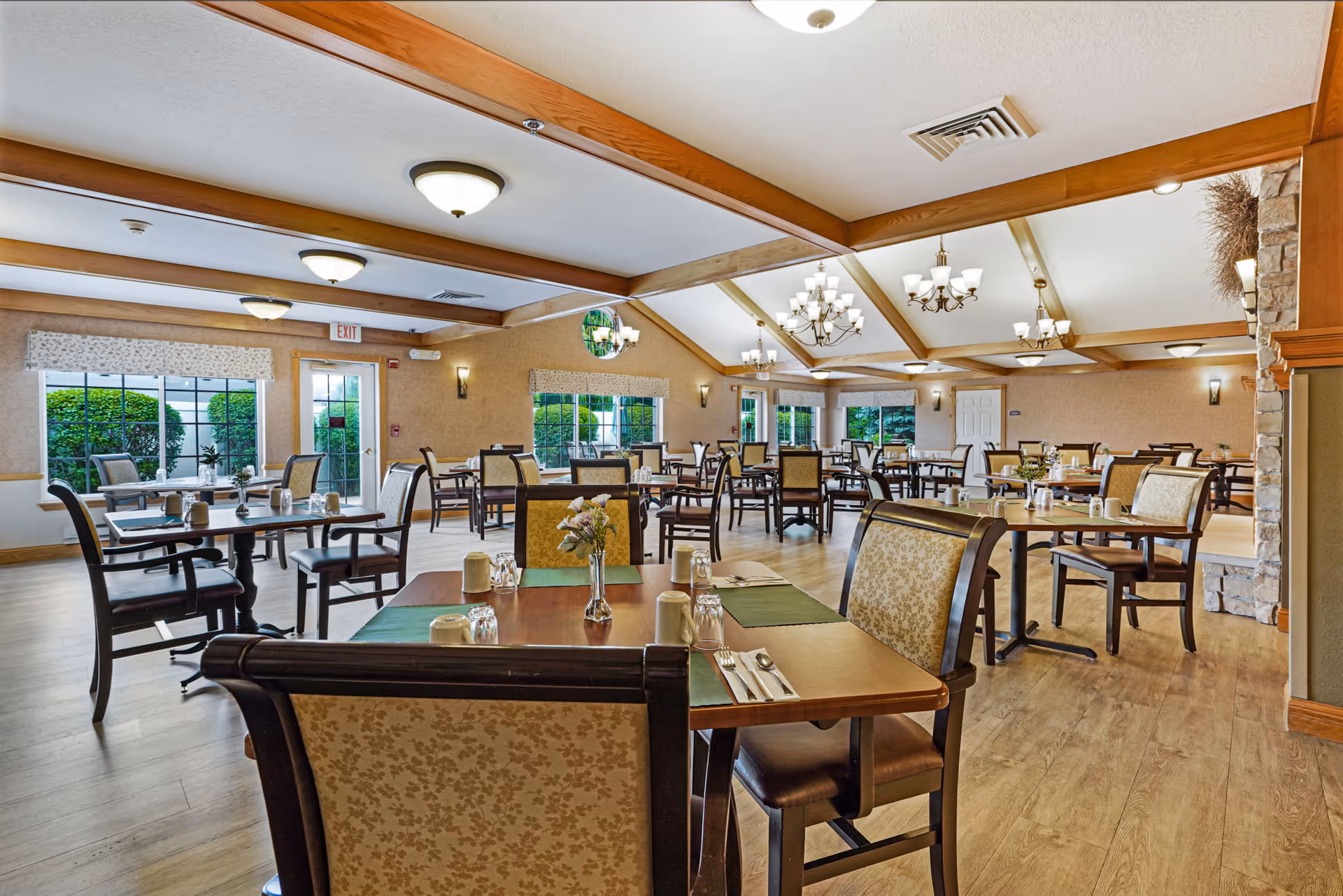 Bright, spacious dining room with multiple tables set for meals, upholstered chairs, chandeliers, and large windows.