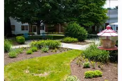 A landscaped outdoor garden area with green grass, various shrubs, and plants. There is a paved walkway winding through the garden, and a red bird feeder is visible on the right side. The exterior of a building with windows and a door is partially visible in the background.