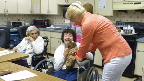 Two elderly women sitting in wheelchairs in a kitchen or dining area, one holding a small brown dog and smiling, while a woman in an orange shirt and white pants bends down to interact with the dog. Another person is partially visible in the background near the kitchen counter.