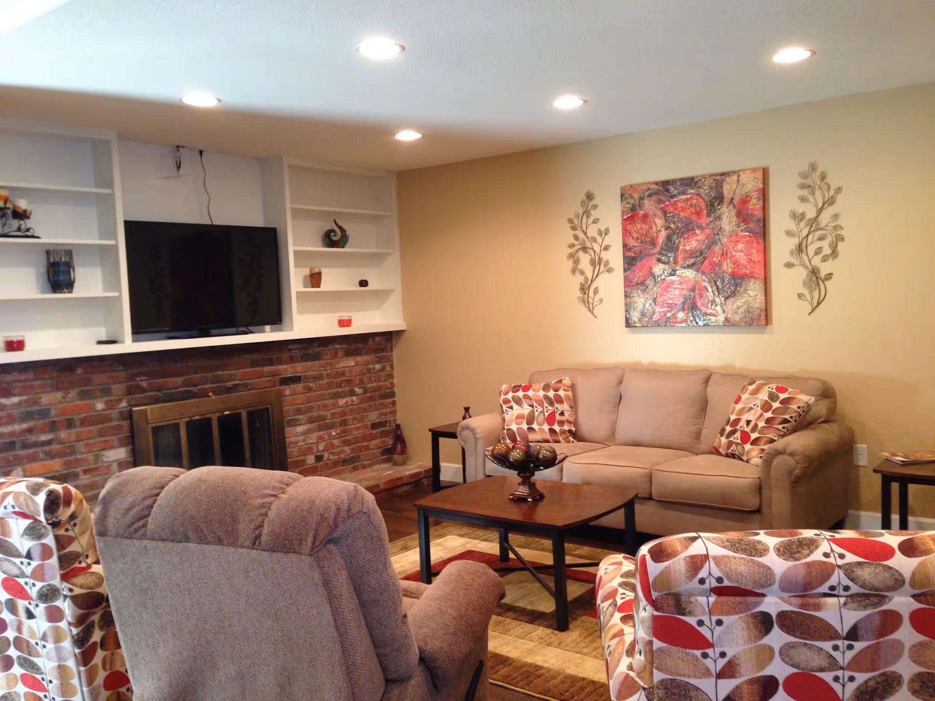 A cozy living room with a beige sofa adorned with patterned cushions, two armchairs with leaf-patterned upholstery, a brown recliner, a wooden coffee table with decorative items, a brick fireplace with a brass screen, built-in white shelves with small decorative pieces, a wall-mounted flat-screen TV, and a colorful abstract painting flanked by metal leaf wall art on a beige wall.