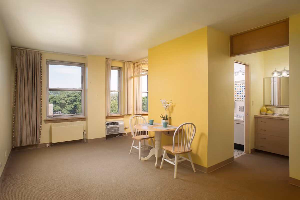 A small dining area in a retirement community apartment with a round wooden table and two chairs, set against a yellow wall. The room has beige carpeting, two large windows with curtains showing a view of trees outside, and an air conditioning unit below one window. To the right, there is an open doorway leading to a bathroom with a vanity, mirror, and light fixtures.