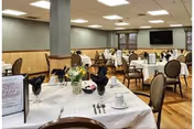 A spacious dining room with multiple tables set for a meal, featuring white tablecloths, black napkins, silverware, cups, and small floral centerpieces. The room has wooden flooring, beige walls with wooden paneling, and a large flat-screen TV mounted on the far wall.