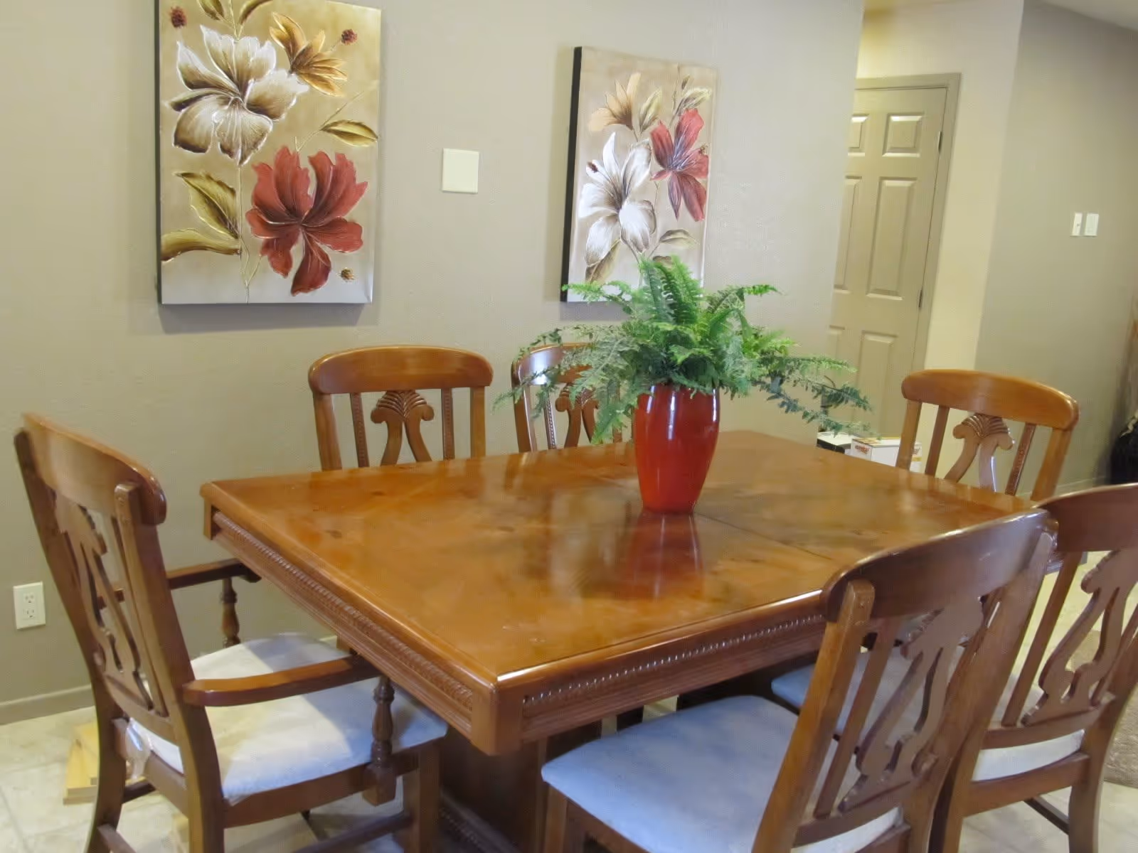 Wooden dining table with six chairs and a red vase holding a fern, with floral paintings on the wall behind.