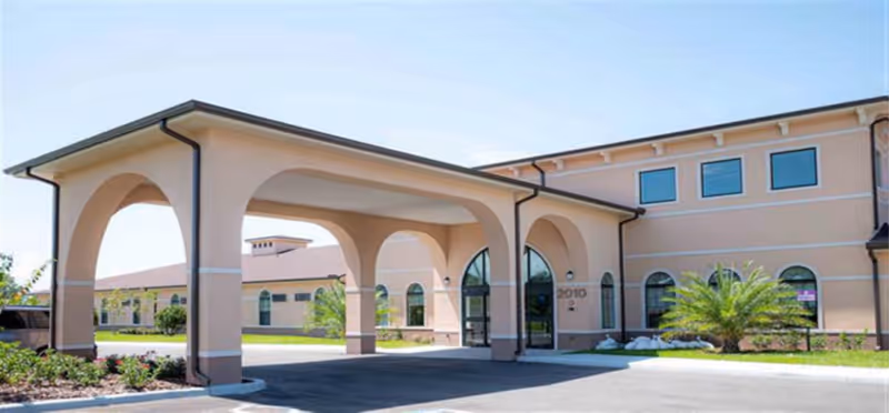 Exterior view of Brilliance Assisted Living facility showing a beige building with arched entryway, large windows, and landscaped greenery including a palm plant near the entrance.
