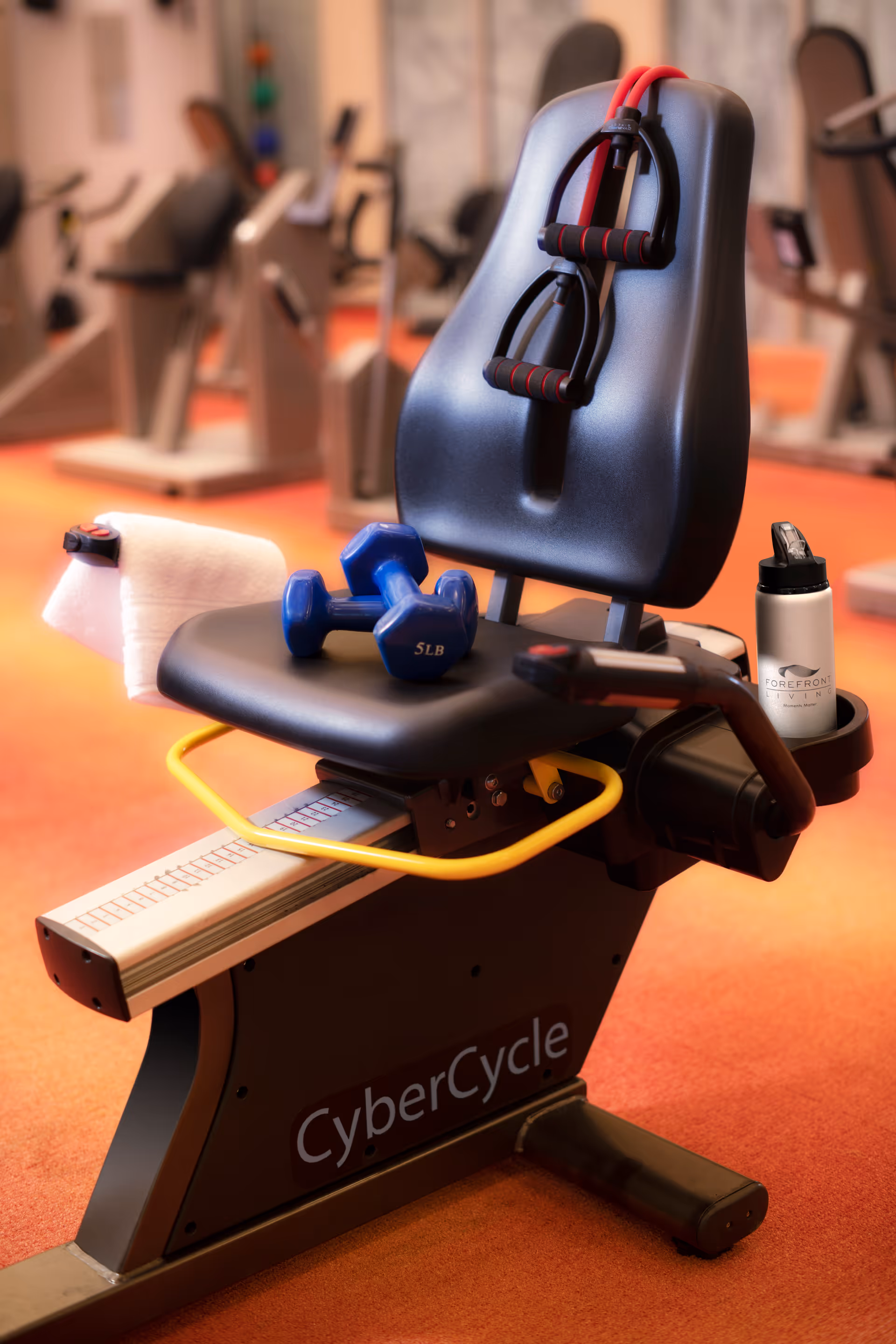 A close-up view of a CyberCycle exercise machine in a gym setting. On the seat are two blue 5-pound dumbbells and a red resistance band with handles hanging over the backrest. A white towel is draped over the side, and a water bottle labeled 'Forefront Living' is placed in the machine's cup holder. The background shows other exercise equipment on an orange carpeted floor.