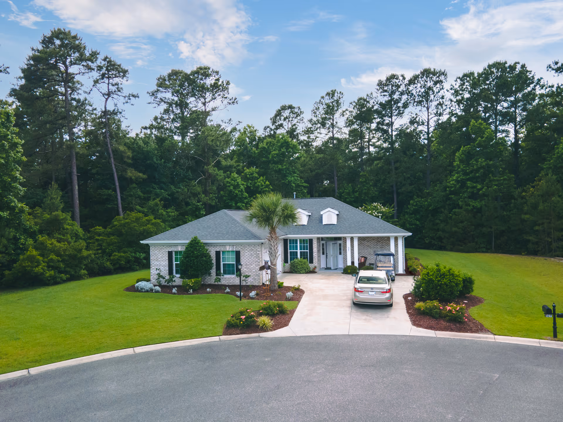 A single-story brick house with a gray roof situated at the end of a cul-de-sac. The house has a driveway with a parked car and a golf cart. The front yard is well-maintained with green grass, shrubs, and a palm tree. Tall trees and dense greenery surround the property under a partly cloudy blue sky.