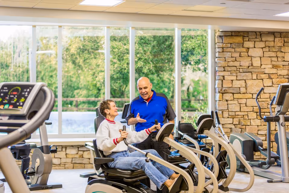 An elderly woman is exercising on a recumbent bike in a fitness room while a man in a blue and black shirt talks to her. The room has large windows with a view of greenery outside and a stone accent wall. Various exercise machines are visible around them.
