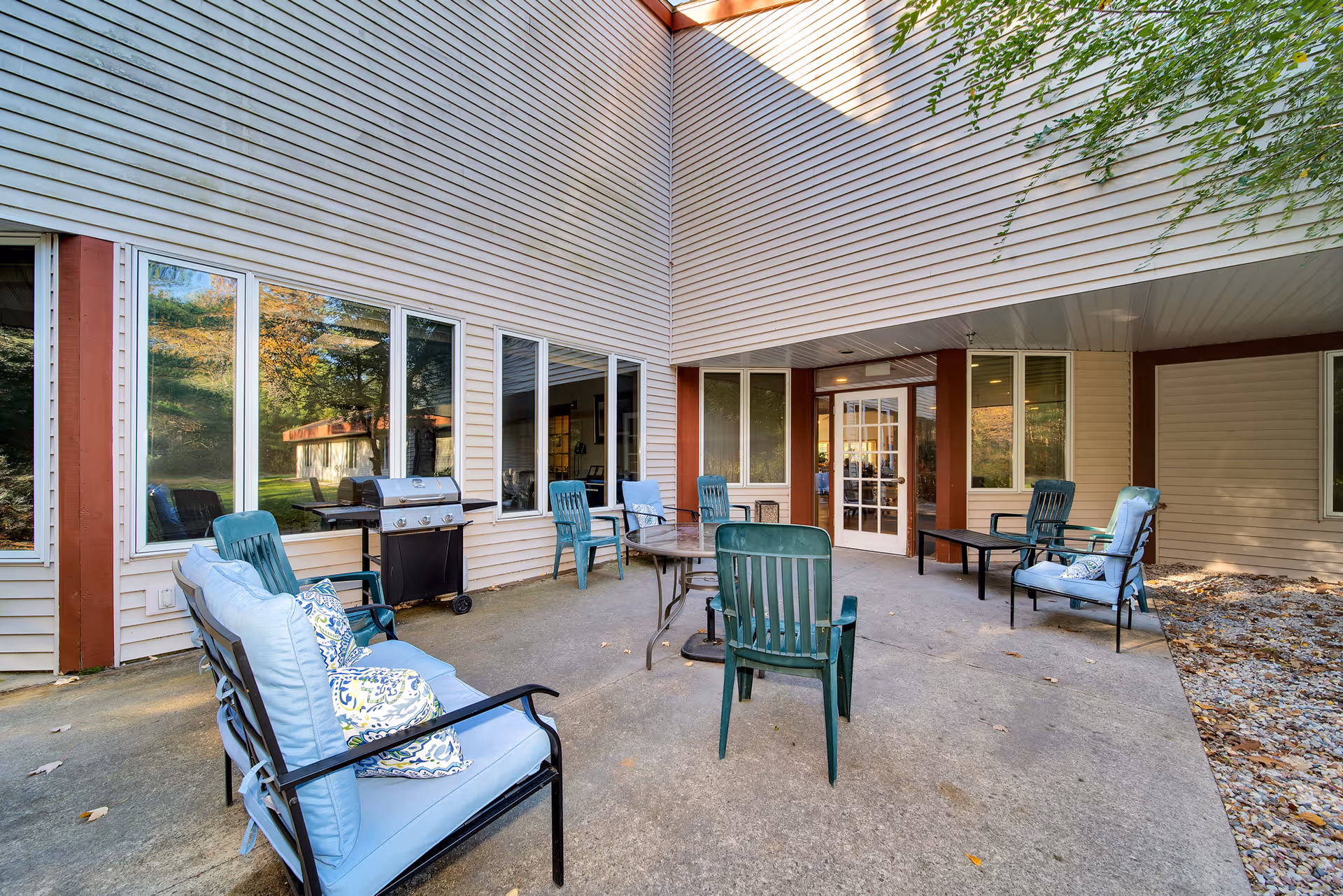 Outdoor patio area at Wellington Place at Biron with several cushioned chairs and a table arranged on a concrete surface. There is a barbecue grill near the windows of the building, which has beige siding and large windows. The area is partially shaded by the building structure.