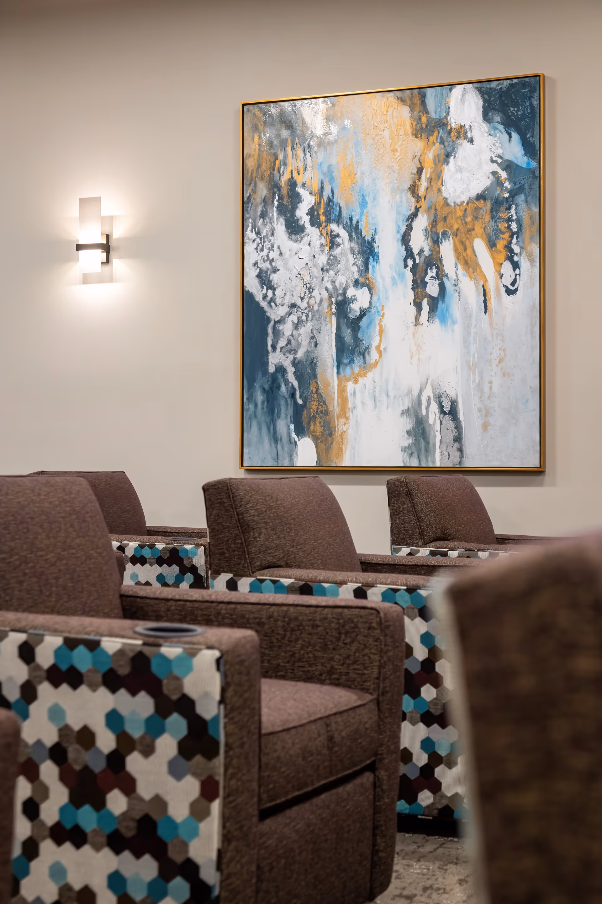 A seating area with brown armchairs featuring hexagonal patterned fabric on the sides in shades of blue, black, and white. A large abstract painting with blue, white, gray, and gold colors hangs on the beige wall above the chairs. A modern wall sconce light fixture is mounted on the wall to the left of the painting.