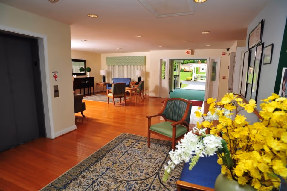 Interior view of a senior living facility lobby area with wooden floors, a patterned rug, green cushioned chairs, a table with yellow and white flowers, and an elevator on the left. In the background, there are more chairs and tables near a window with blinds, and a glass door leading outside.