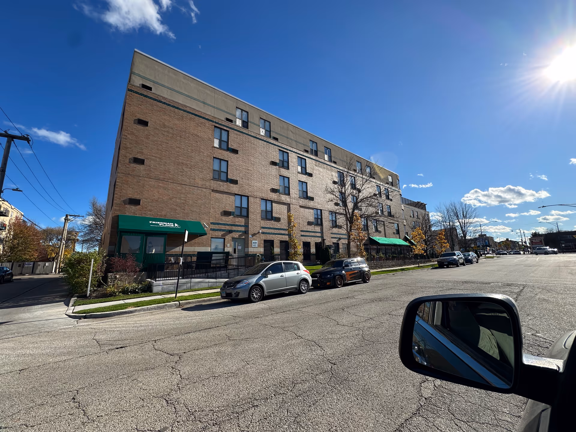 Exterior view of a multi-story brick building with green awnings labeled Friedman Place. Several cars are parked along the street in front of the building under a clear blue sky with the sun shining brightly.