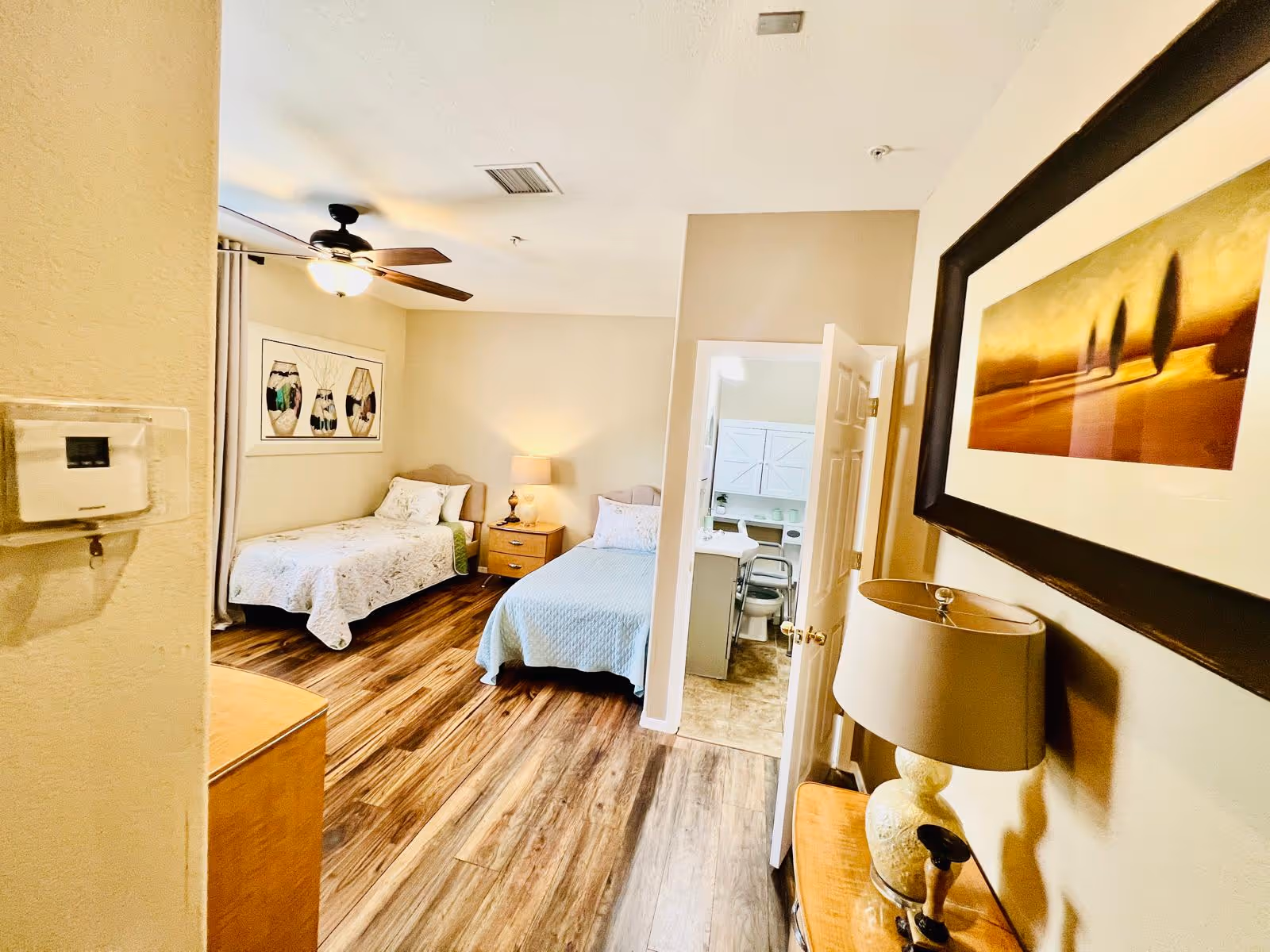 A bright bedroom with two single beds, each with a quilted bedspread, a wooden nightstand with a lamp between them, a ceiling fan with light, and a framed artwork on the wall. The room has wood flooring and beige walls. An open door reveals a small adjoining bathroom with a sink, mirror, and toilet.