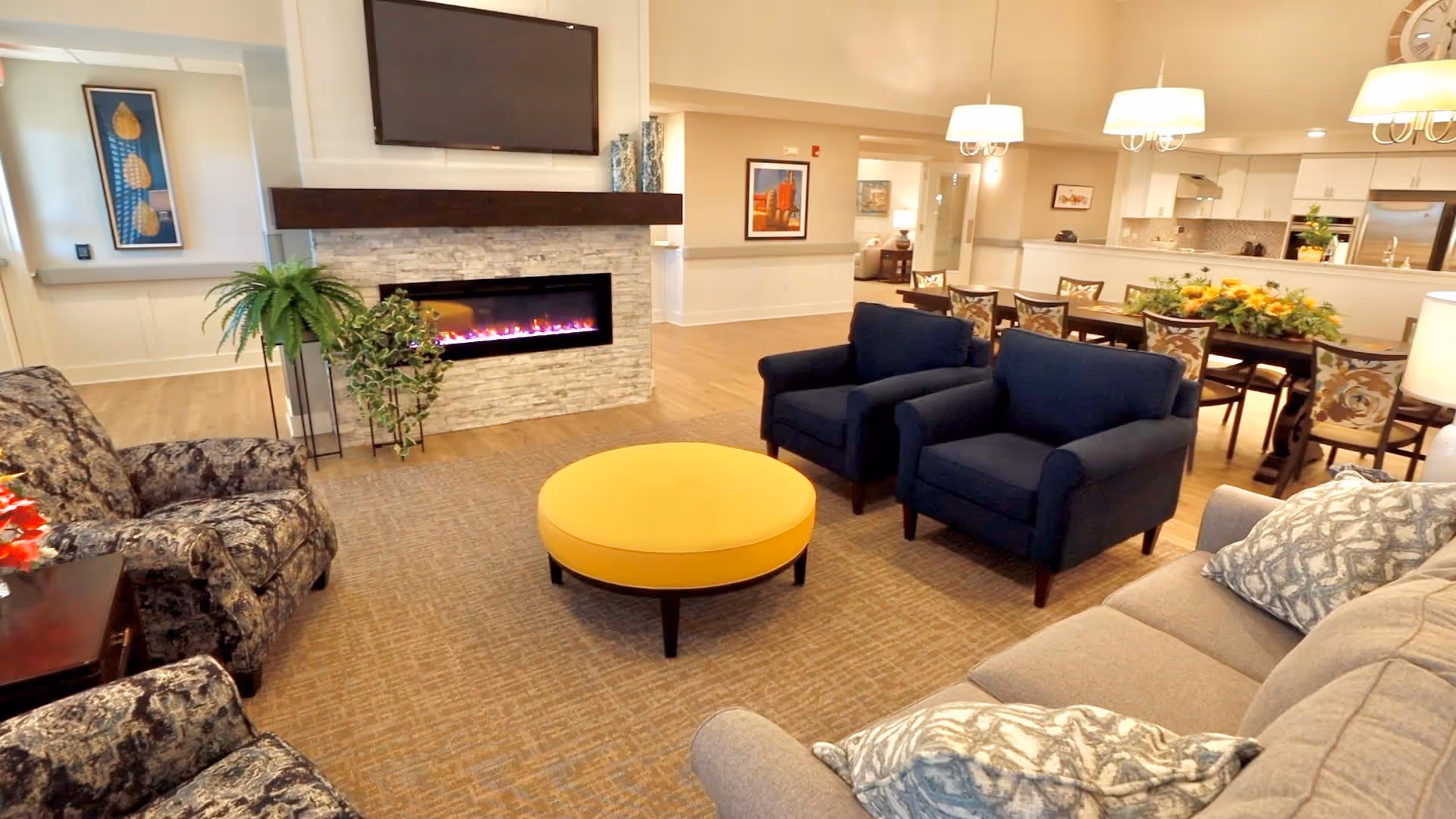 A cozy living room area in an assisted living facility featuring a stone fireplace with an electric fire, a mounted flat-screen TV above it, two blue armchairs, a round yellow ottoman, a patterned armchair, and a beige sofa with patterned pillows. In the background, there is a dining area with a long table, floral cushioned chairs, hanging pendant lights, and a kitchen with white cabinetry and stainless steel appliances.