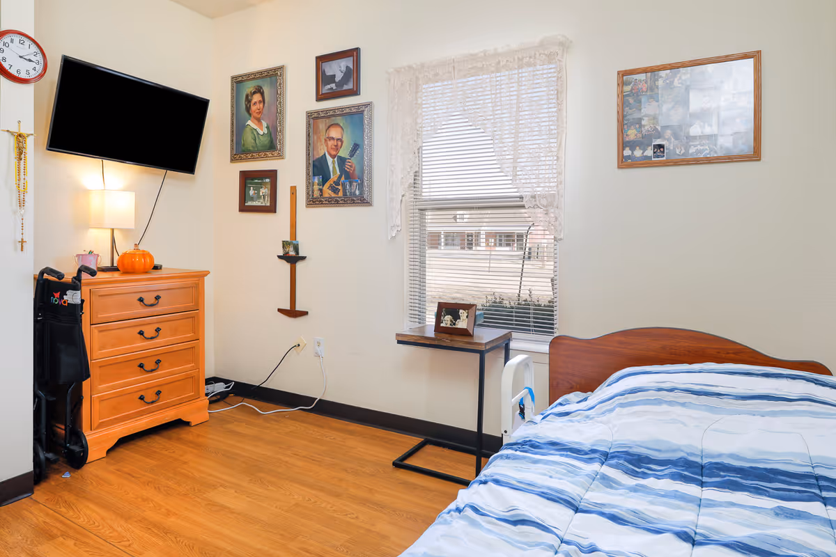 A cozy bedroom in a senior living facility featuring a single bed with blue and white striped bedding, a wooden dresser with a lamp and a small pumpkin on top, a wall-mounted flat-screen TV, and several framed pictures on the walls. There is a window with blinds and lace curtains letting in natural light, and a small table beneath the window with a framed photo. A walker is positioned next to the dresser.