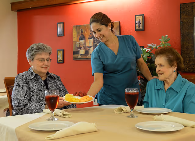 A caregiver serves a plate of fruit to two elderly women seated at a dining table.