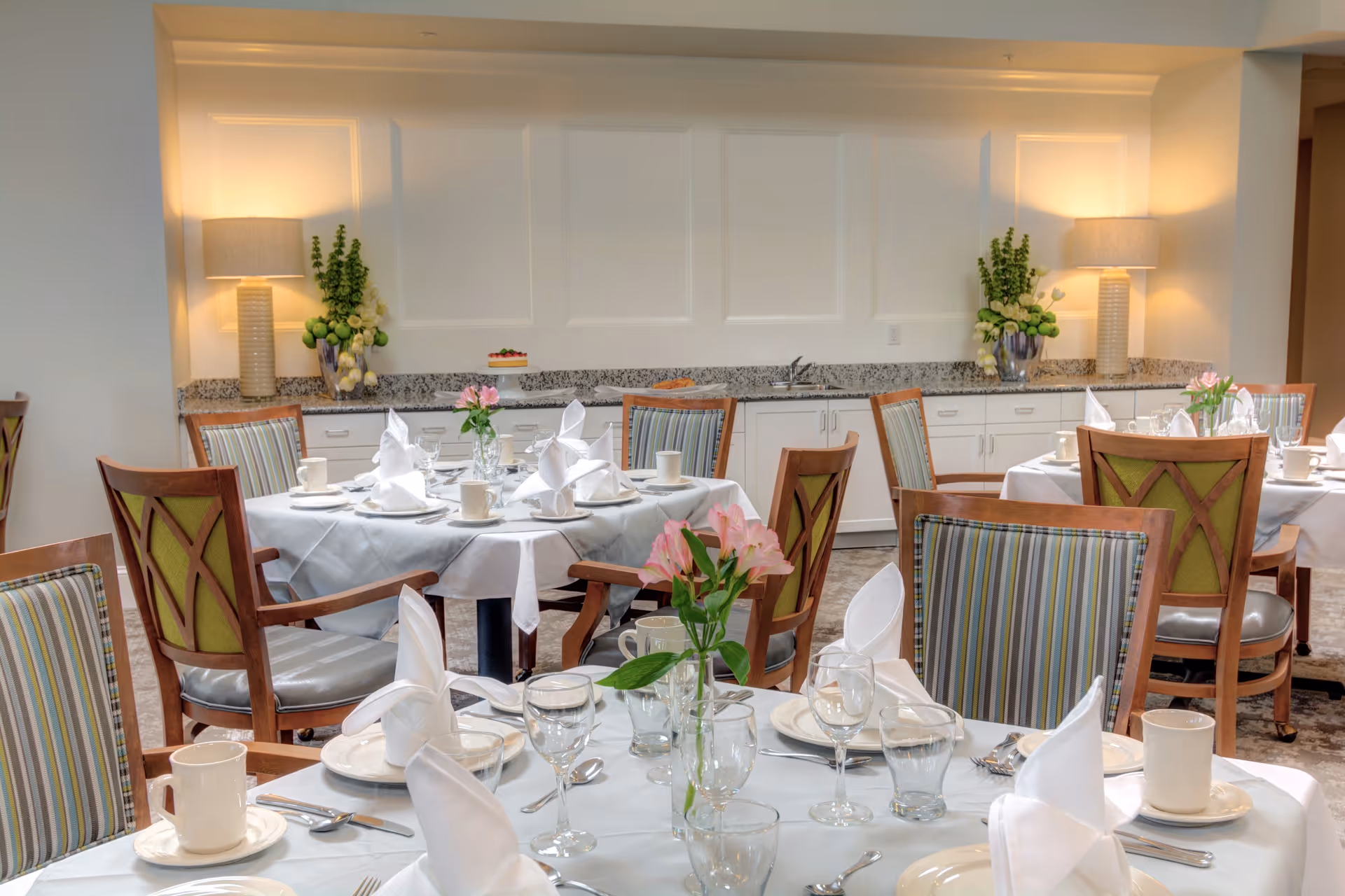 A dining room in a senior living facility with round tables covered in white tablecloths, set with white napkins, plates, cups, and glassware. Each table has a small vase with pink flowers. The room features wooden chairs with striped and green upholstery. In the background, there is a granite countertop with decorative plants and two lamps providing warm lighting.