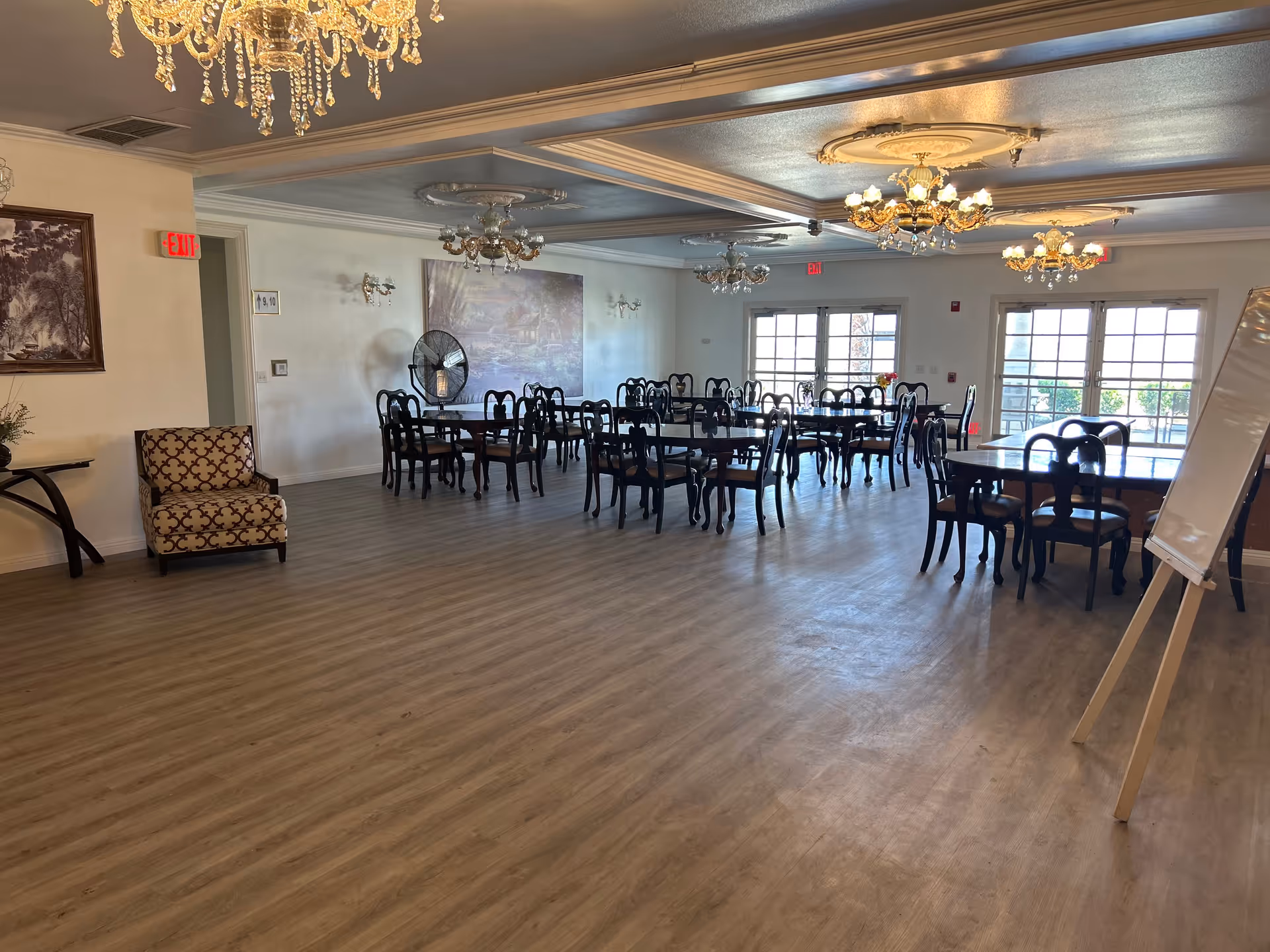 A spacious dining room with multiple dark wooden tables and chairs arranged neatly. The room features elegant chandeliers hanging from the ceiling, large windows and glass doors letting in natural light, a patterned armchair near the wall, a standing fan, and a whiteboard on an easel. The floor has a light wood finish and the walls are decorated with paintings and sconces.
