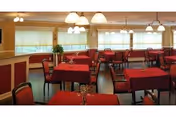 Dining room with multiple tables covered in red tablecloths and chairs beneath hanging light fixtures and windows with shades.