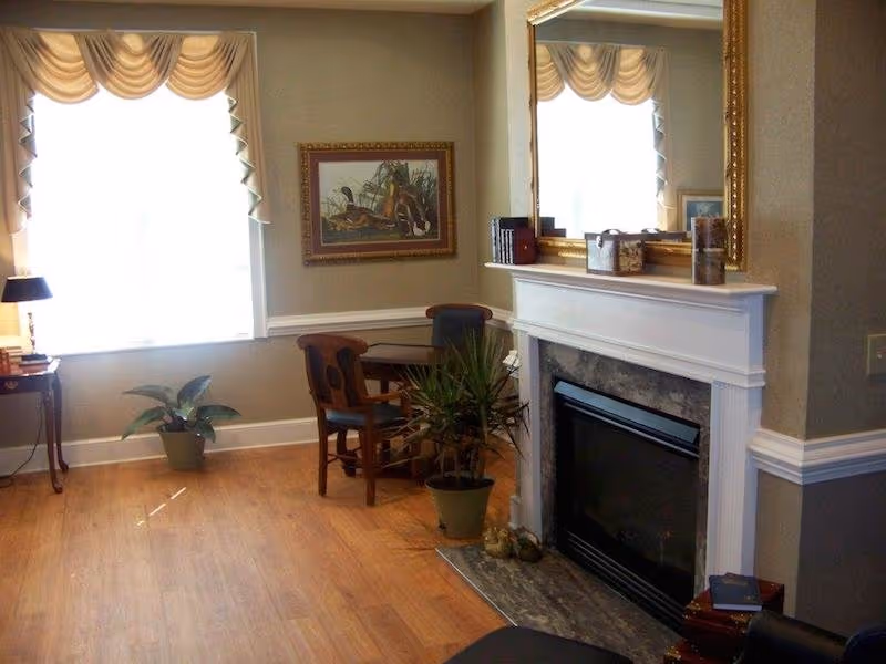 A cozy living room area with a fireplace featuring a white mantel and a large gold-framed mirror above it. There is a small wooden table with two chairs, a potted plant on the floor, and a window with beige curtains letting in natural light. A framed painting hangs on the wall above the table.