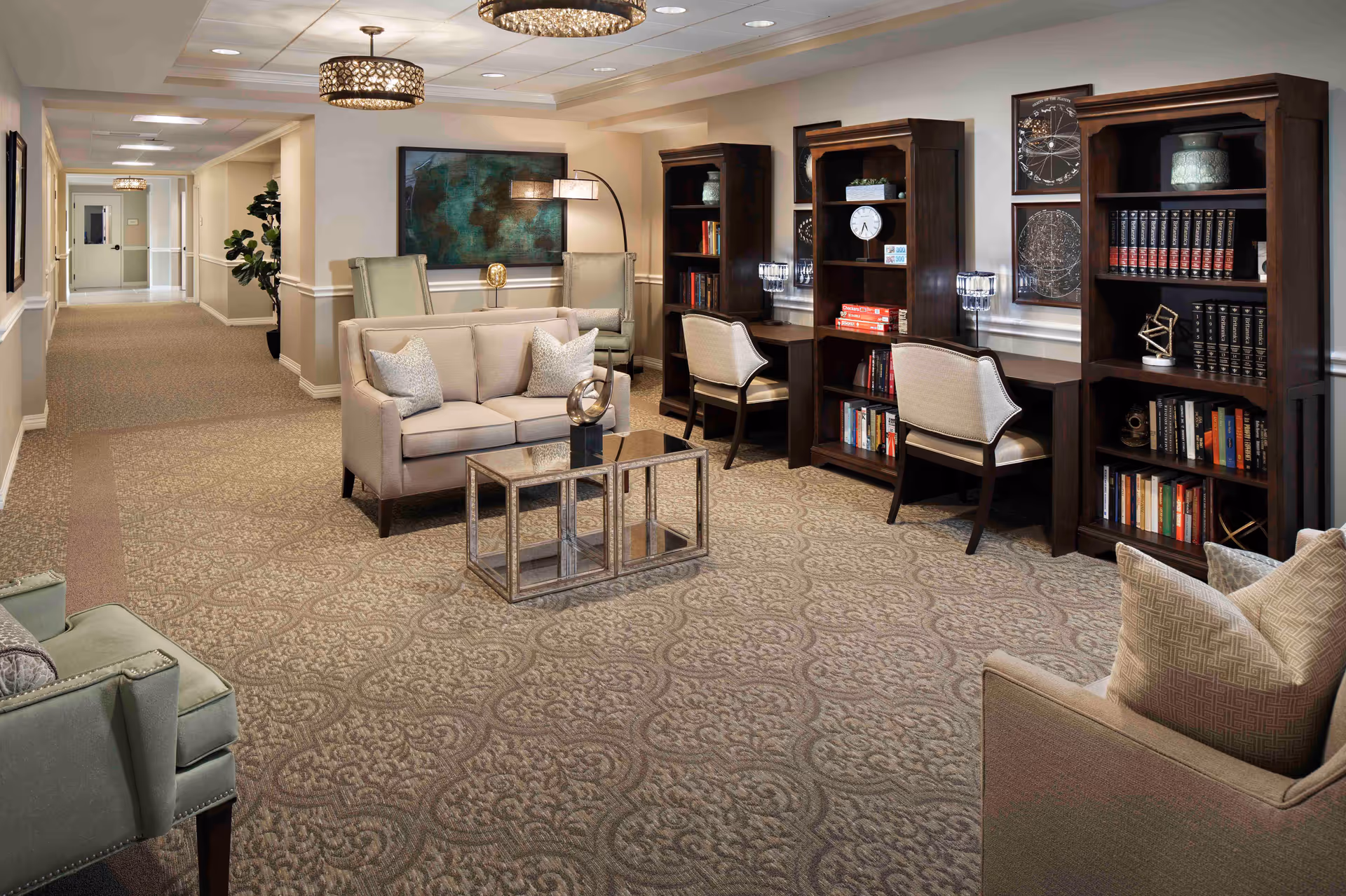 Well-appointed senior living common area with sofas, armchairs, bookshelves and study desks along a carpeted hallway.
