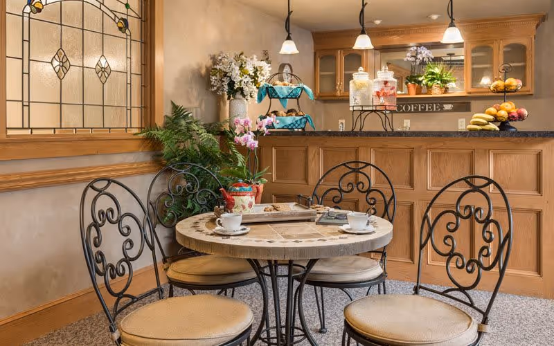 A cozy seating area with a round table and four wrought iron chairs with beige cushions. On the table are two white coffee cups, a tray with a teapot, and some reading materials. Behind the table is a wooden counter with glass jars, a tiered tray with snacks, and a sign that says 'COFFEE'. The background includes wooden cabinets with glass doors, pendant lights, and decorative plants. A stained glass window is visible on the left wall.