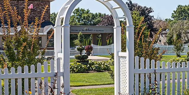 White wooden archway and picket fence leading to a green lawn with landscaped bushes and trees. In the background, a sign reads 'Welcome to The Arbors at Hauppauge' with a building partially visible behind it under a clear blue sky.