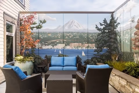 Outdoor patio area with four dark wicker chairs with blue cushions arranged around a matching square table. Behind the seating area is a large mural depicting a scenic view of a lake with sailboats, trees, buildings, and a snow-capped mountain in the background. There are potted plants and a stone planter with greenery on the right side.