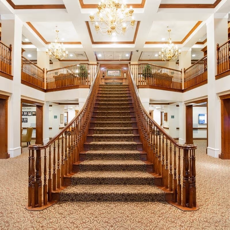 A grand interior space featuring a wide, carpeted staircase with wooden railings leading to an upper level balcony. The area is well-lit with multiple ornate chandeliers hanging from a coffered ceiling. The walls and columns are white, and the flooring is carpeted with a patterned design. There are doorways and seating areas visible on the ground floor.