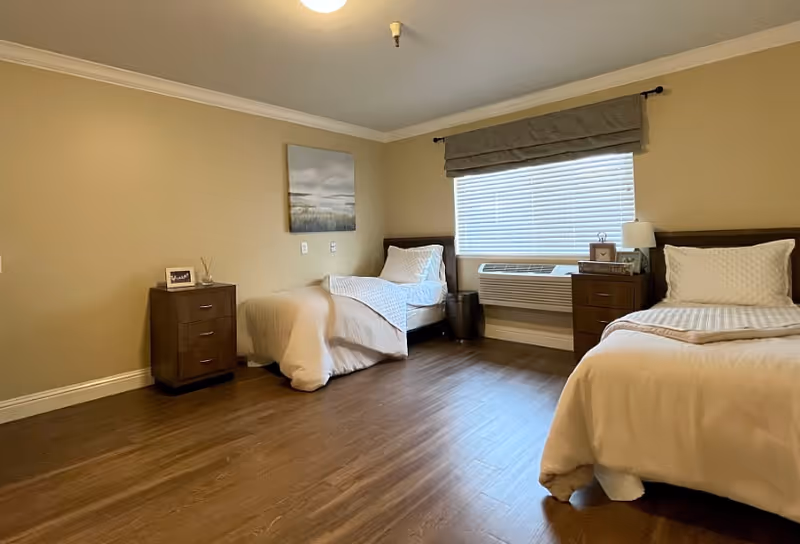 Spacious bedroom with two twin beds, nightstands, a window with blinds and an air-conditioning unit.