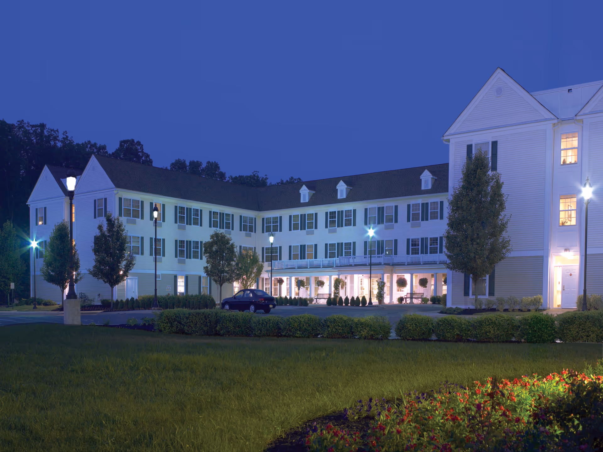 Exterior view of a large, three-story senior living facility building at dusk with lights on inside and outside. The building has white siding, multiple windows with green shutters, and a well-maintained lawn and landscaping in the foreground. Several street lamps illuminate the parking area and entrance.