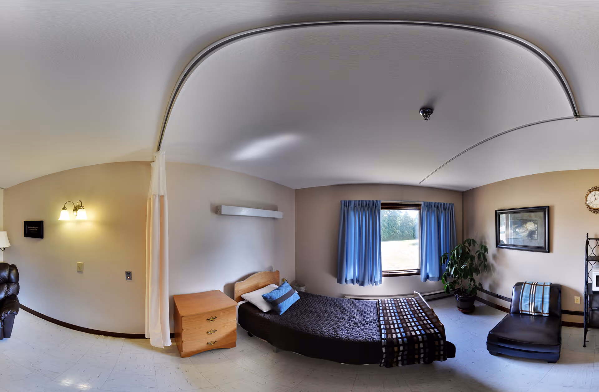 A senior living facility bedroom with a single bed covered in a dark quilt and pillows, a wooden nightstand with drawers, a window with blue curtains letting in natural light, a potted plant, a framed picture on the wall, a clock, and a cushioned chair with a striped pillow. The room has a light-colored floor and walls, and a ceiling track for a privacy curtain.
