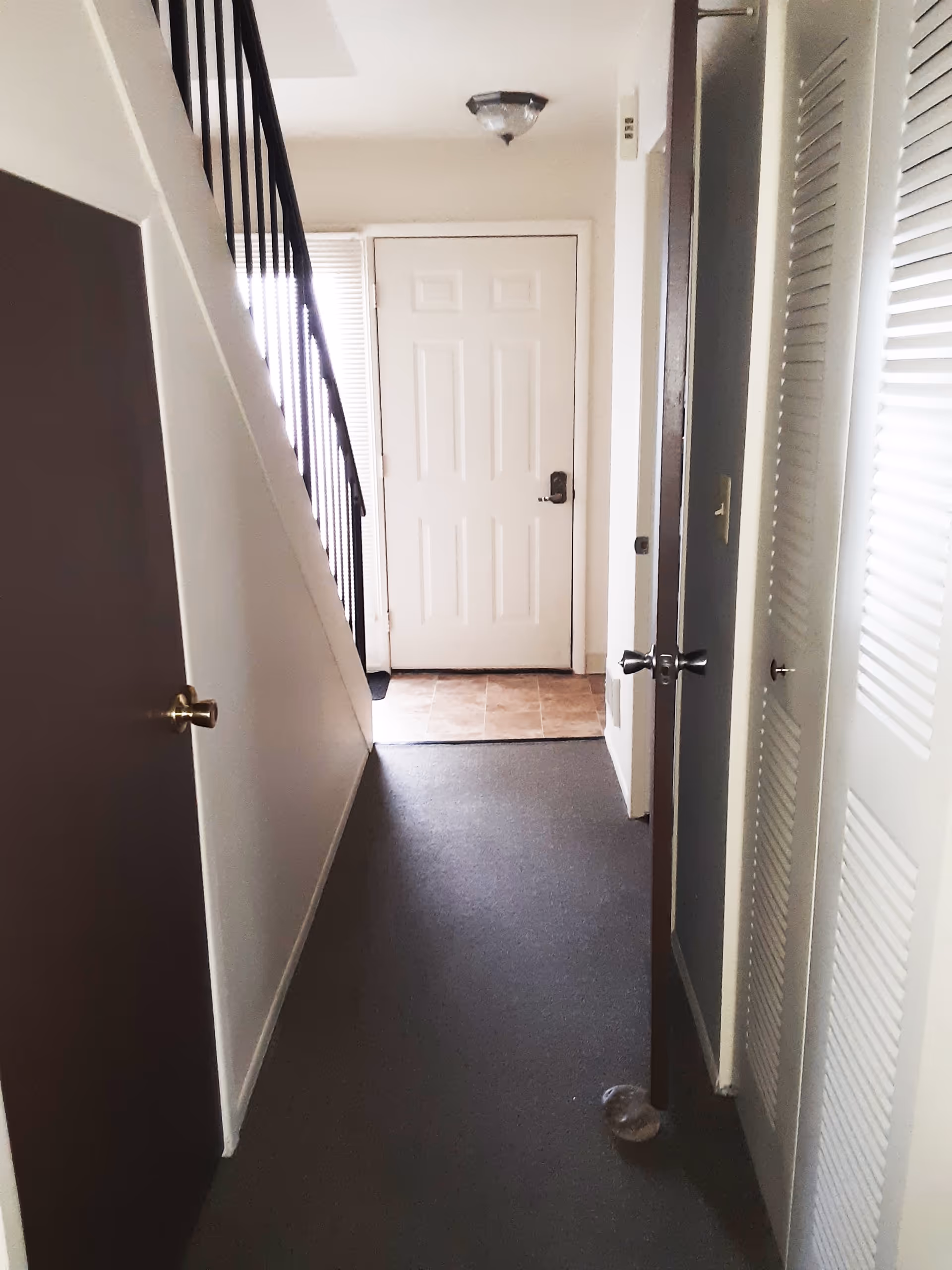 Interior hallway of a facility with a white front door at the end, a staircase with black railing on the left, and several doors on the right side. The floor is carpeted except near the door where there are tiles. A small object is on the floor near one of the doors.