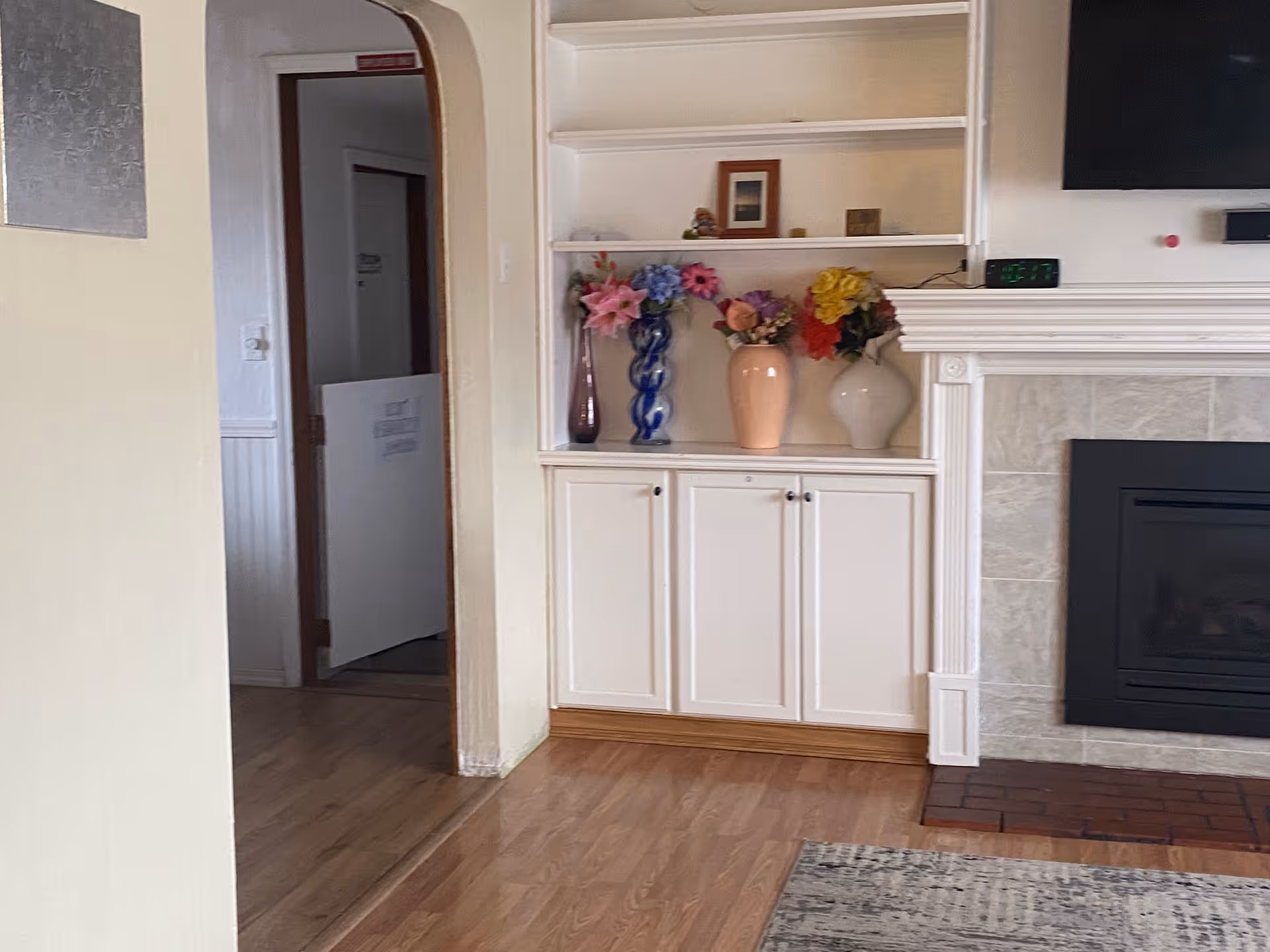 Interior view of a living room area with a built-in white cabinet and shelves displaying vases with colorful flowers. To the right is a fireplace with a tiled surround and a mounted TV above it. The floor is wooden, and there is a doorway with an arched frame leading to another room.