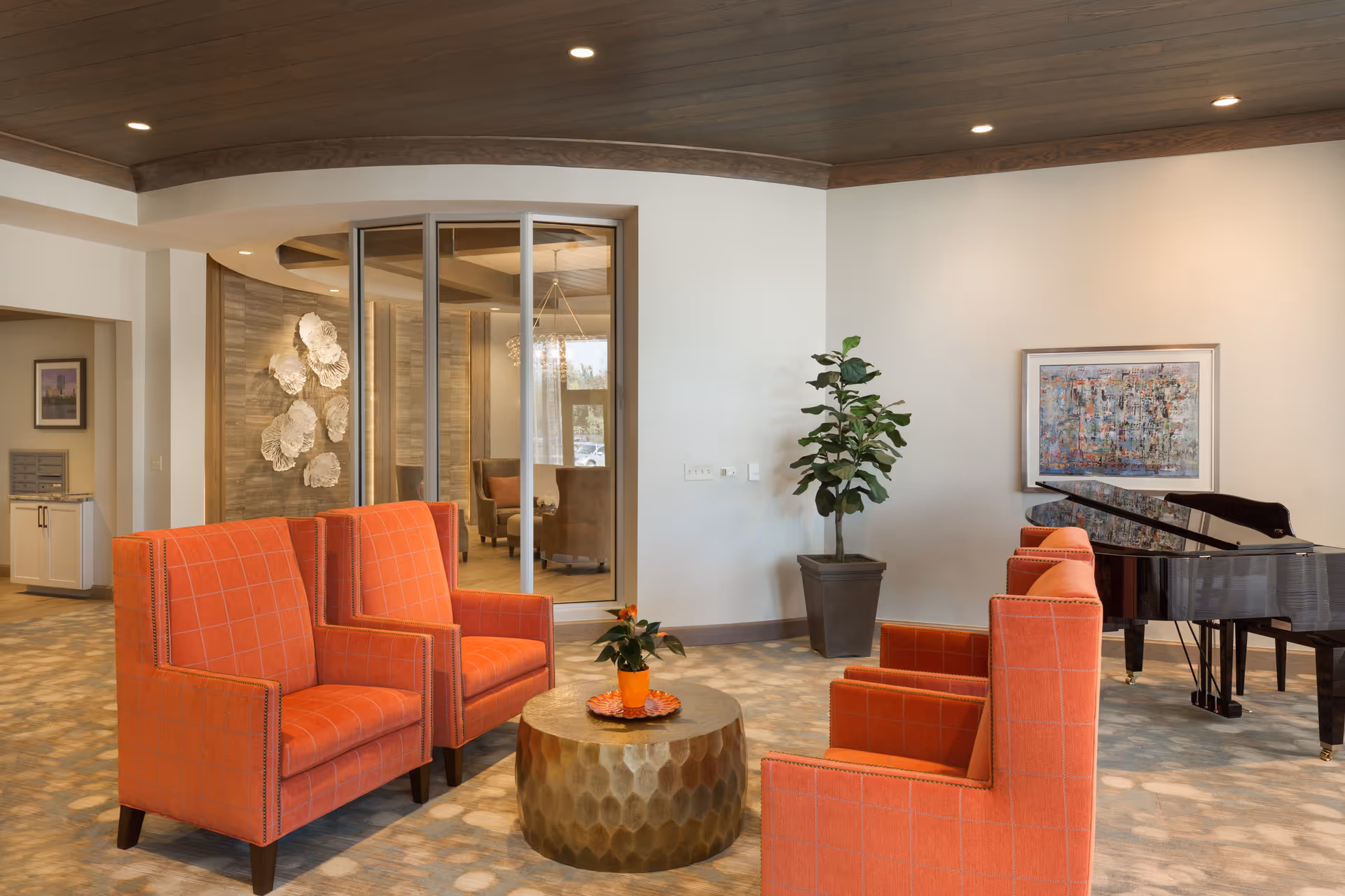 A modern living room area with four orange upholstered armchairs arranged around a round metallic coffee table with a small potted plant on it. The room features a wooden ceiling with recessed lighting, a large potted plant in the corner, a black grand piano, and a colorful abstract painting on the wall. In the background, there is a glass partition revealing another seating area with armchairs and decorative wall art.