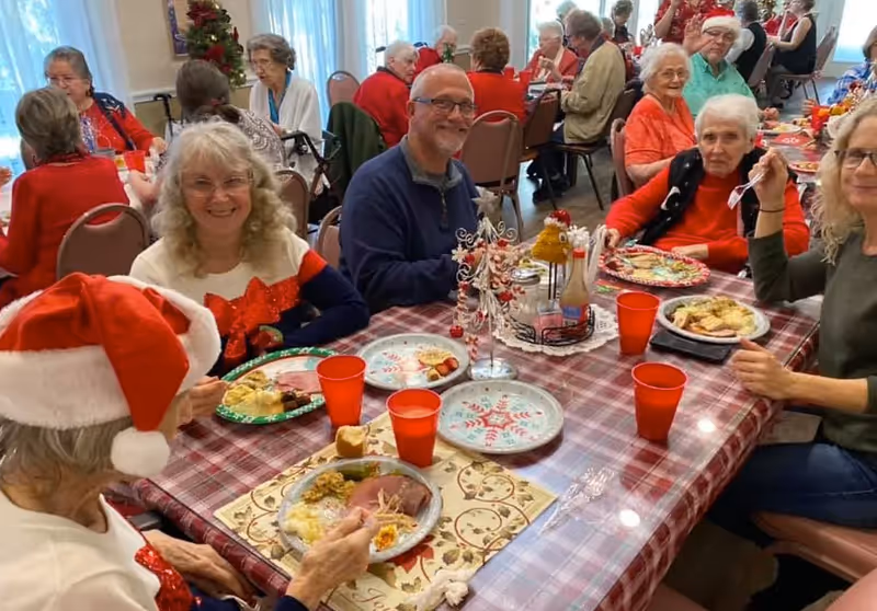 A group of elderly people gathered around a table enjoying a festive meal together. The table is covered with a red and white checkered tablecloth and decorated with holiday-themed plates and a small Christmas tree centerpiece. Some individuals are wearing holiday attire, including a Santa hat. The room is bright with large windows in the background.