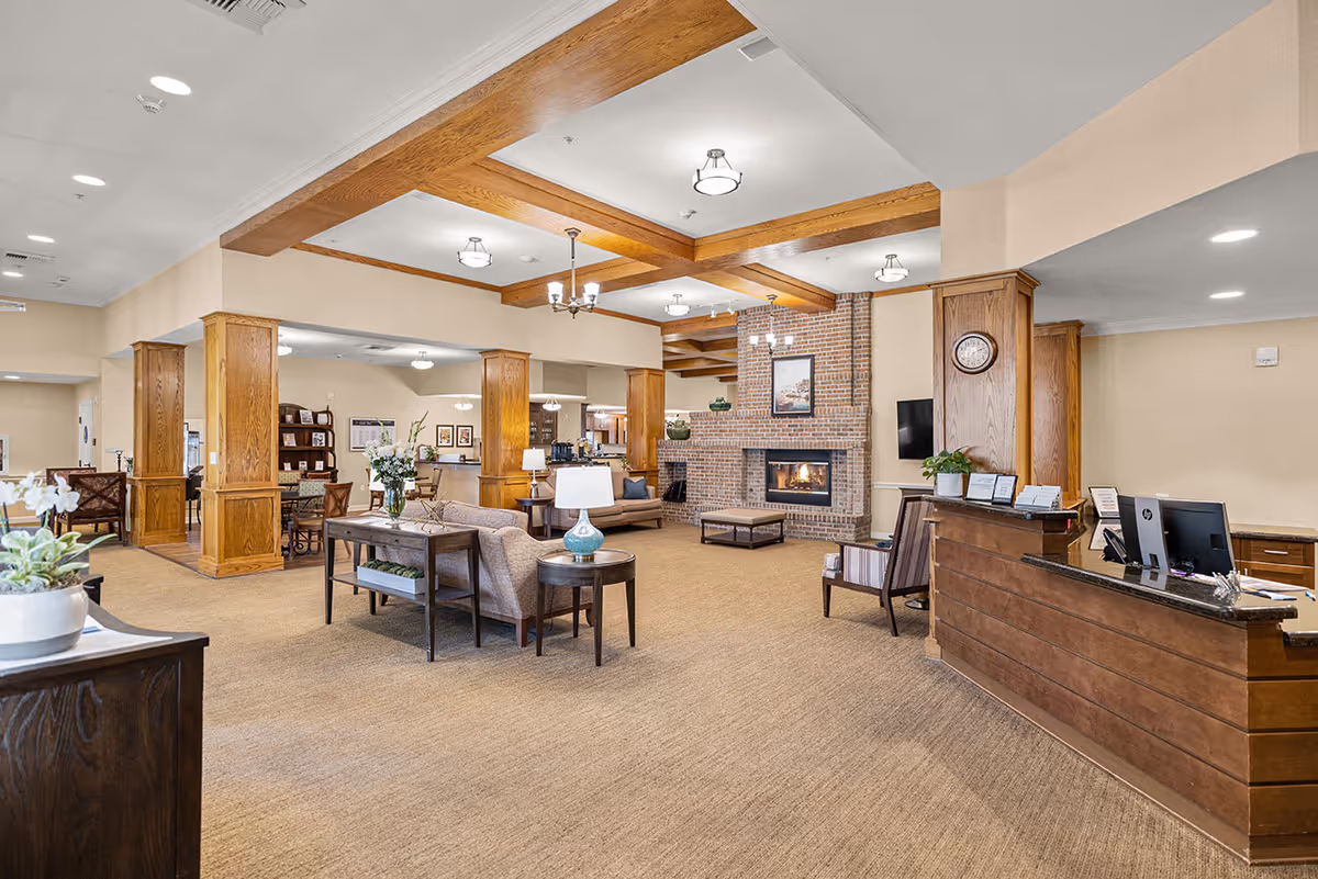 A spacious and well-lit senior living facility common area with beige walls and carpet, wooden ceiling beams, and a brick fireplace. The room features comfortable seating including sofas and chairs, a reception desk with computer monitors, decorative plants, and framed artwork on the walls.