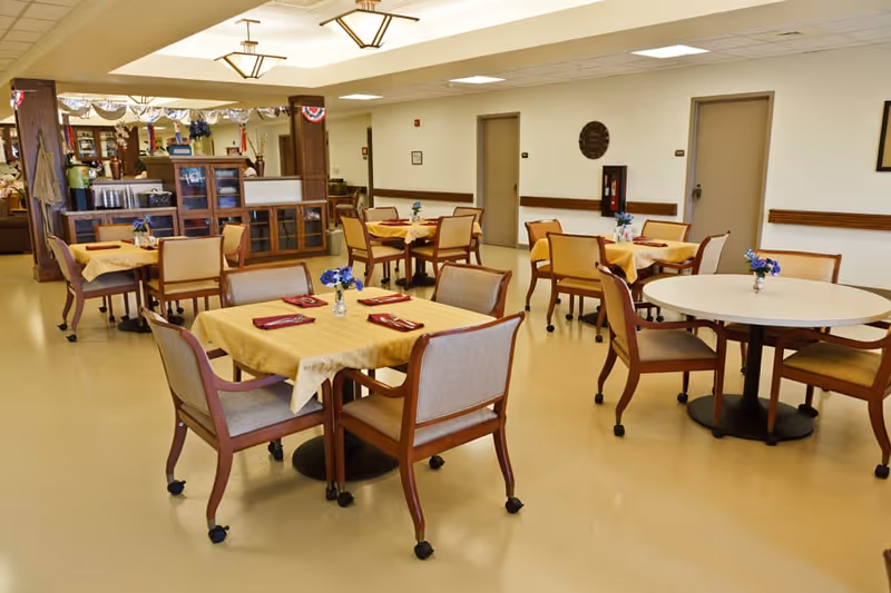 A dining room in a senior living facility with several tables covered with yellow tablecloths, each set with red napkins and small flower vases. The room has beige walls, multiple chairs around each table, and a cabinet with glass doors in the background.