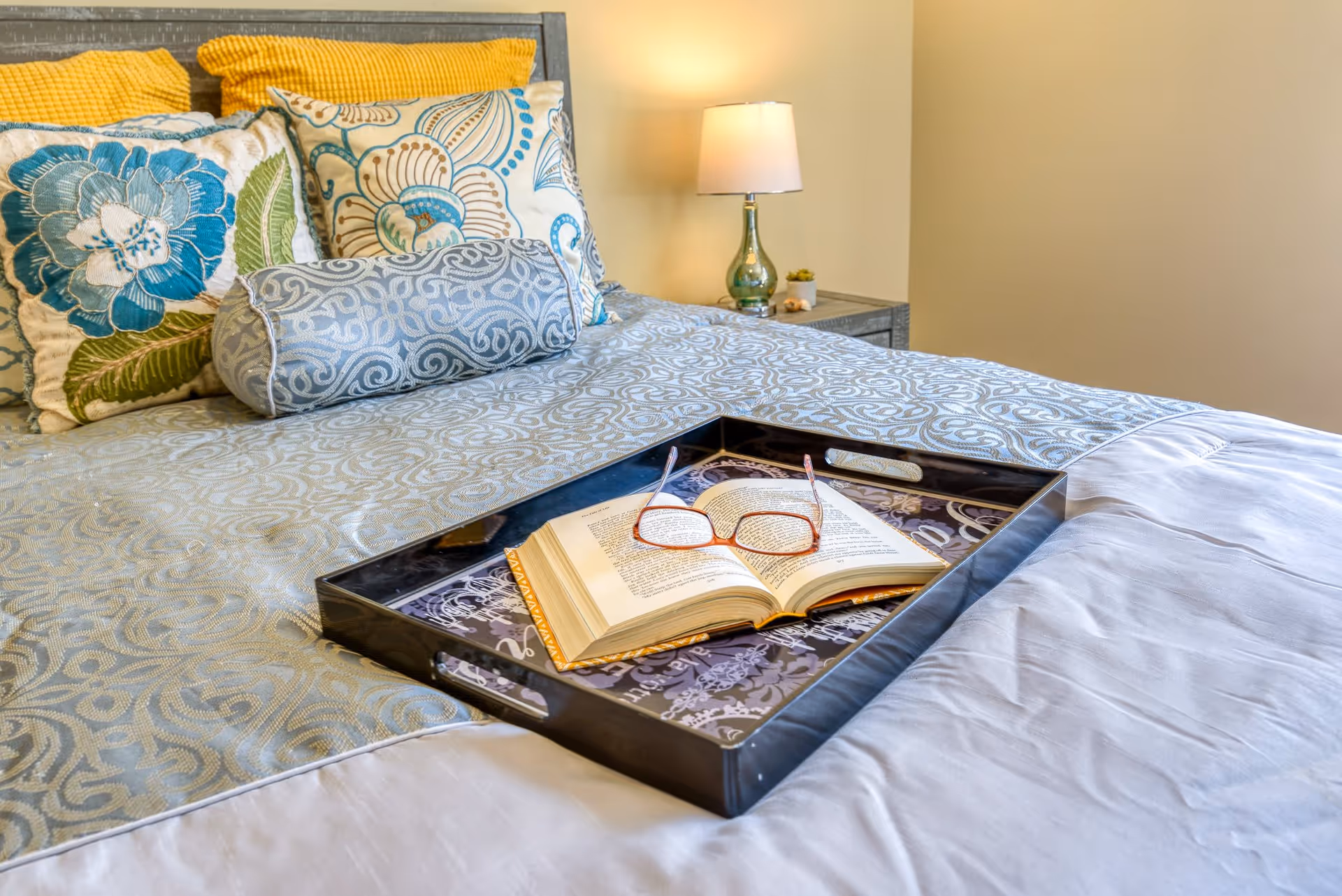 A neatly made bed with decorative pillows, a tray holding an open book and glasses, and a bedside lamp.