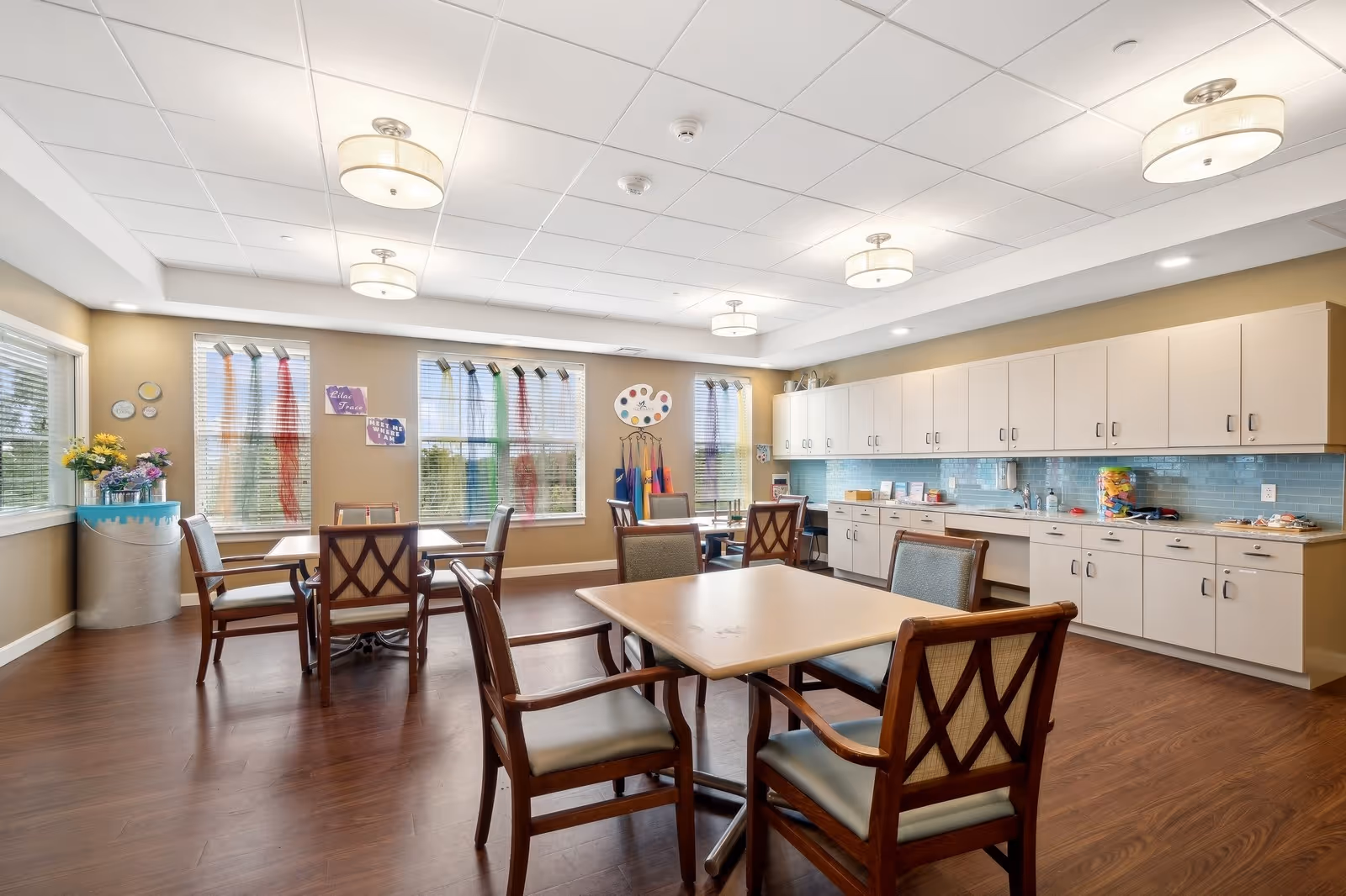 A bright and spacious room with several wooden tables and chairs arranged for group seating. The room features large windows with blinds, allowing natural light to fill the space. Along one wall, there are white cabinets with a blue tiled backsplash and a countertop holding various items. The ceiling has multiple round light fixtures, and the floor is a warm-toned wood. The room appears clean and organized, suitable for dining or group activities.
