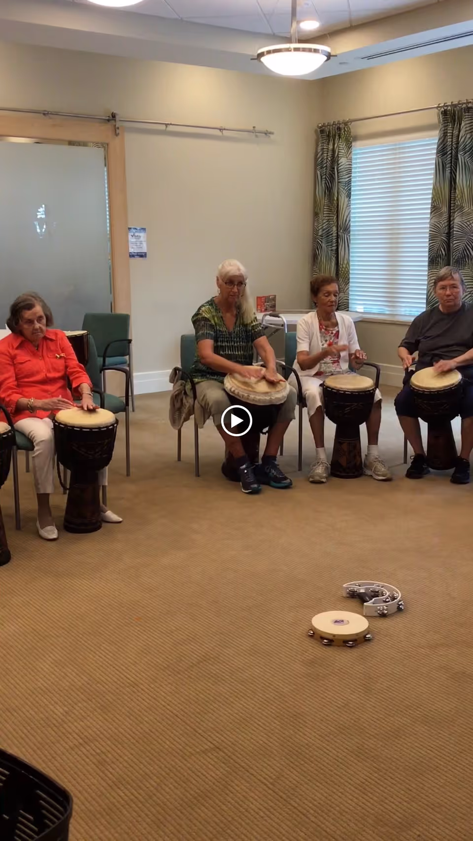Four elderly women sitting in chairs in a room playing hand drums. Two tambourines are placed on the carpeted floor in front of them. The room has beige walls, a window with patterned curtains, and a ceiling light fixture.
