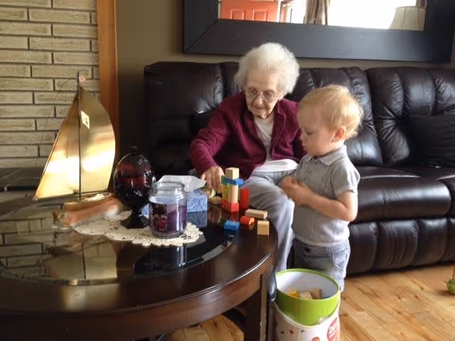 An elderly woman and a young child playing with colorful wooden blocks on a round glass coffee table in a living room with a dark leather couch and a large mirror on the wall behind them.