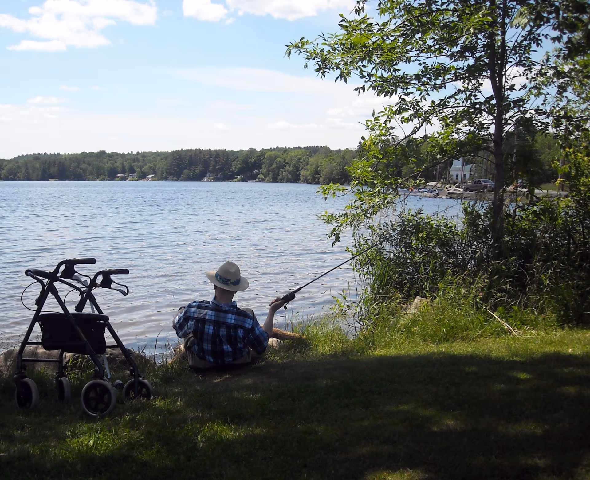 An older person wearing a hat sits on the grassy shore fishing by a lake with a walker beside them.