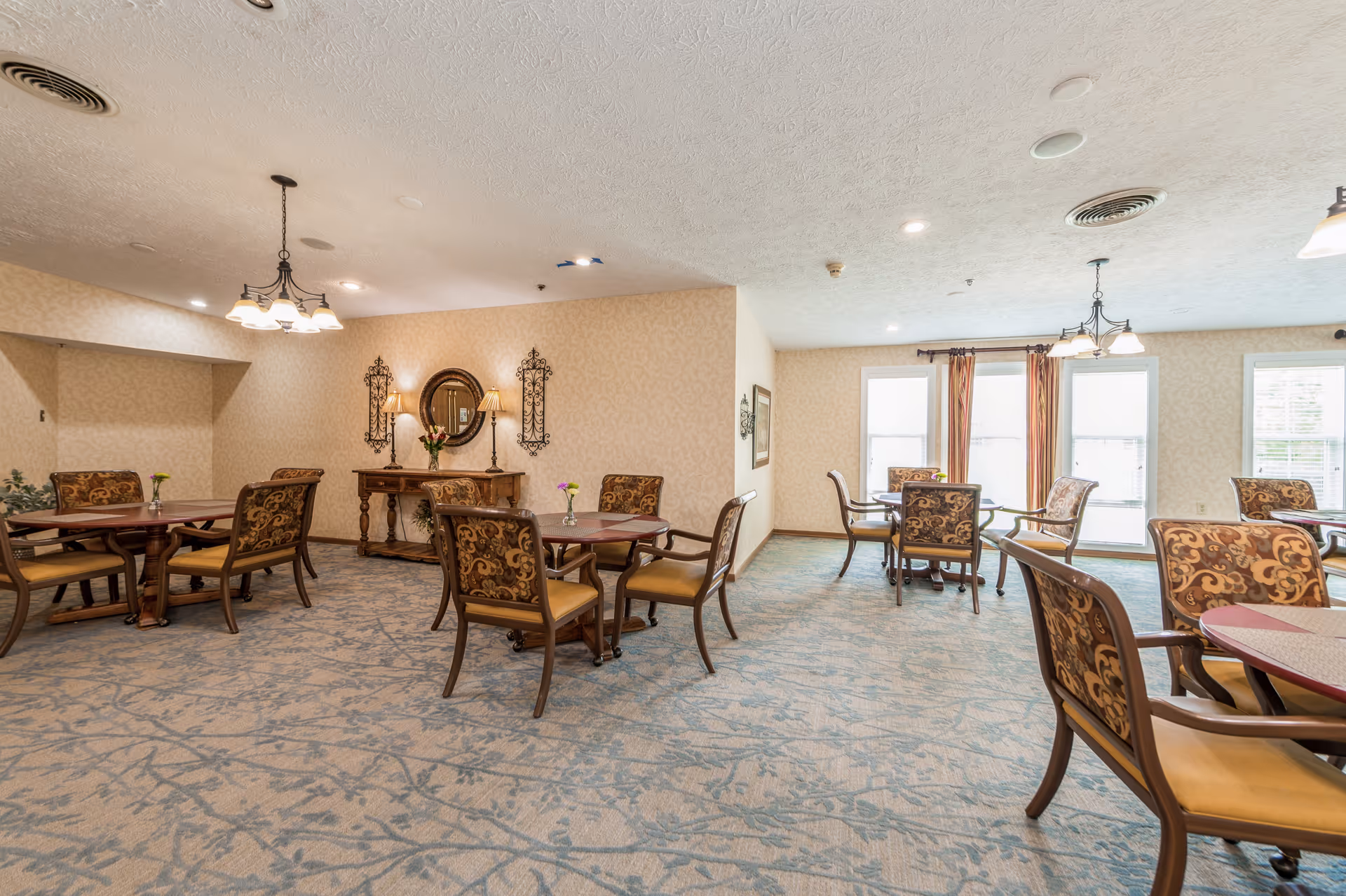 A well-lit dining room with multiple round tables and cushioned chairs featuring patterned upholstery. The room has beige wallpaper, a blue patterned carpet, and decorative wall sconces with a mirror above a wooden console table. Large windows with curtains allow natural light to enter the space.