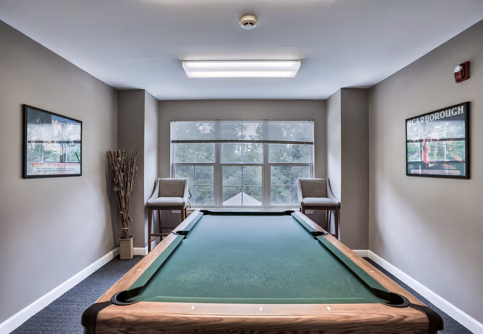 A room with a pool table in the center, two high chairs positioned near a large window with blinds, neutral-colored walls with framed pictures, and a ceiling light fixture.